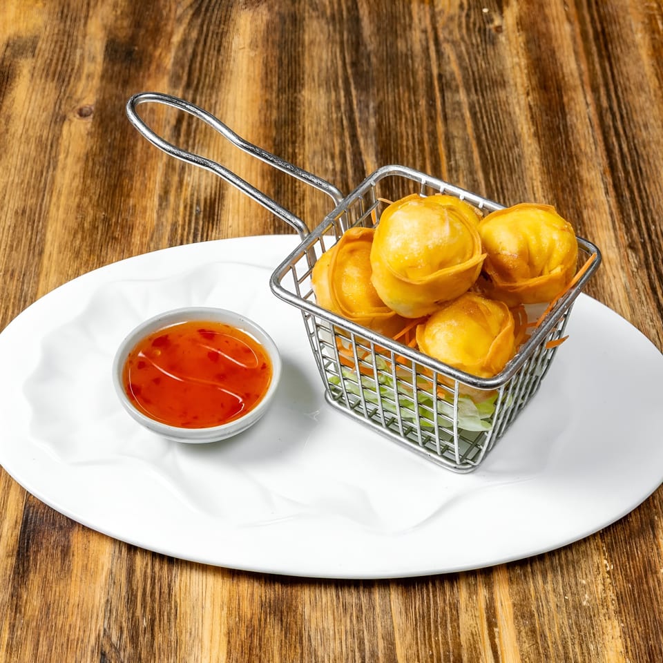 Golden fried wontons in a metal basket with sweet chili sauce on a white plate.