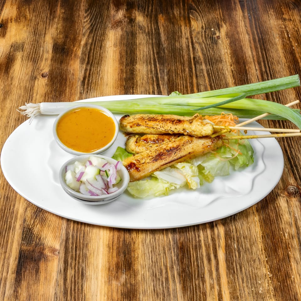 Link Thai chicken satay skewers with peanut sauce, cucumber salad, and green onions on a white plate.