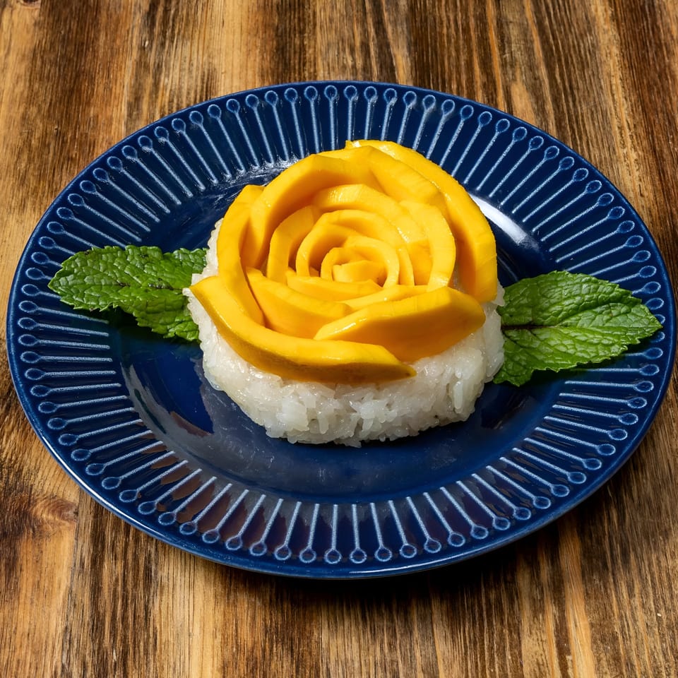 Thai mango sticky rice dessert, with mango slices arranged like a rose, on a blue plate.
