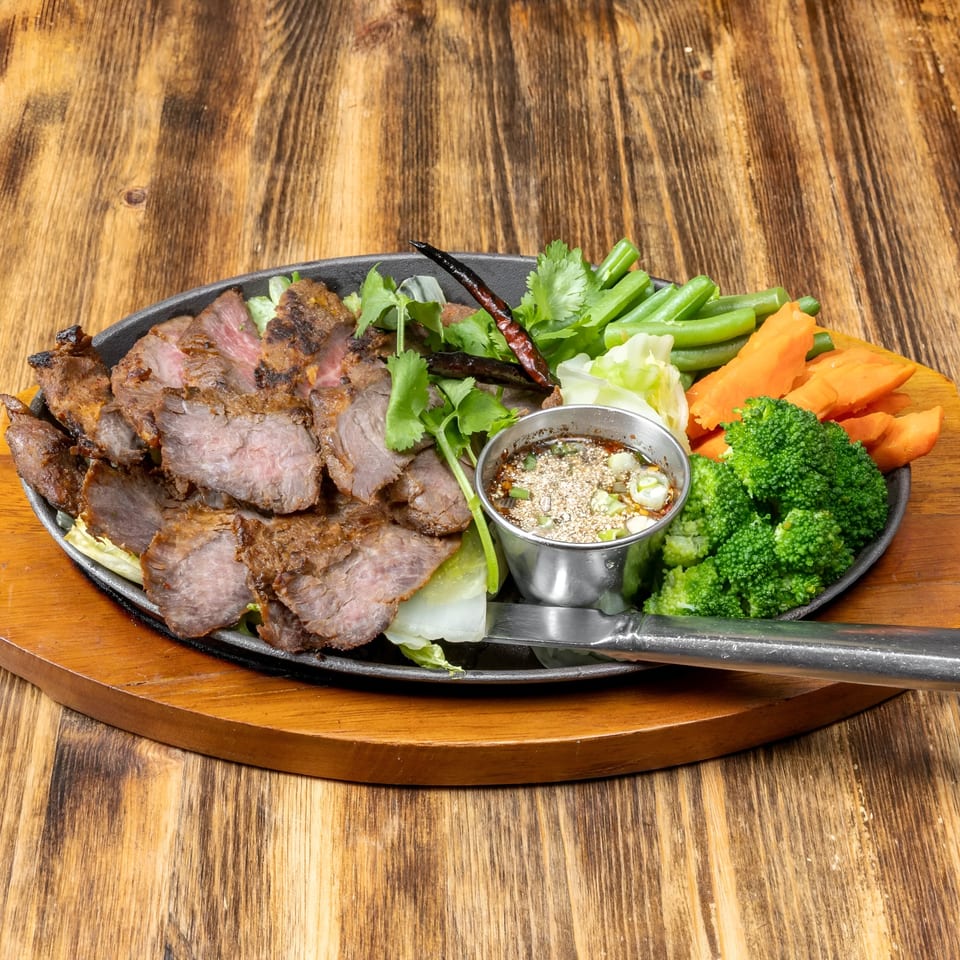 Sliced grilled beef with broccoli, carrots, green beans, and dipping sauce on a wooden table.