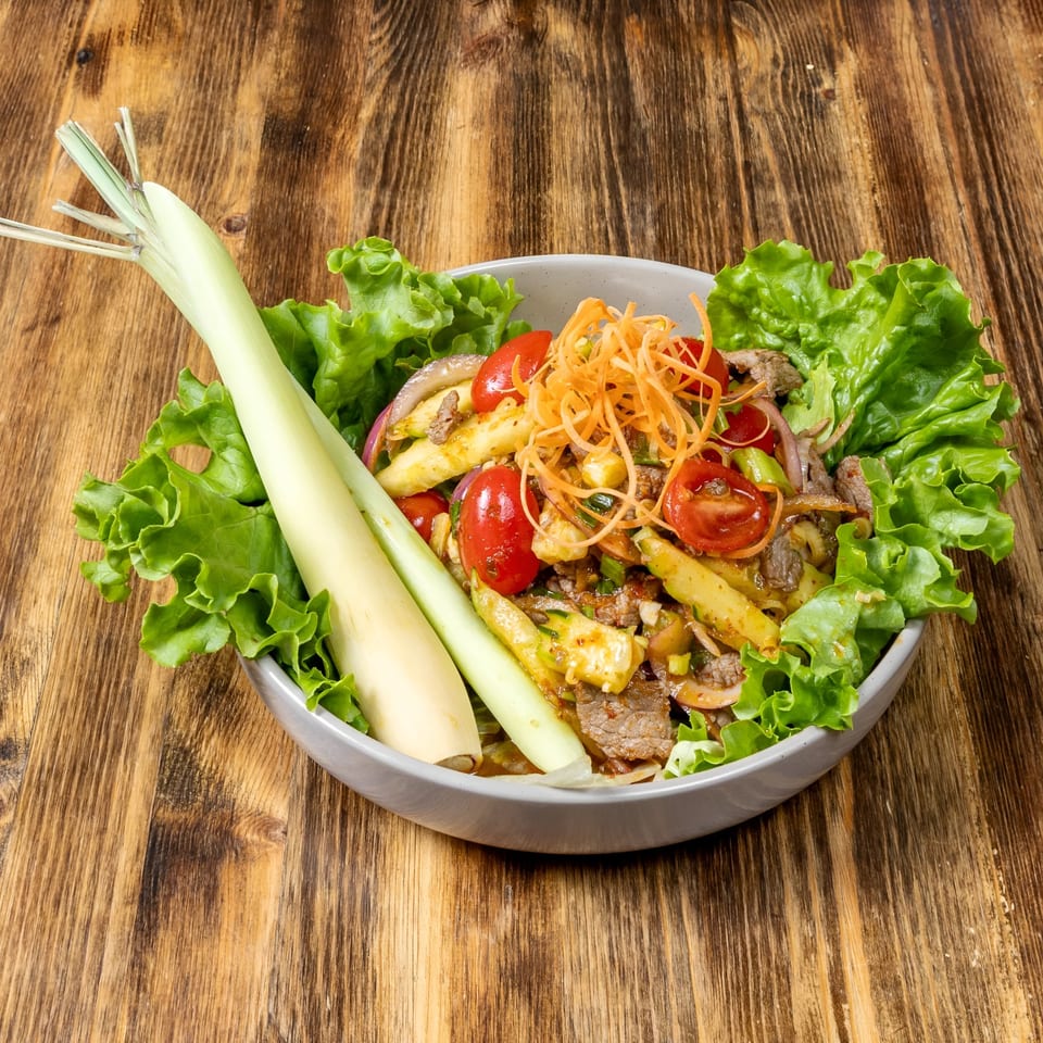 Thai beef salad with lettuce, tomatoes, and lemongrass in a gray bowl on a wooden table.