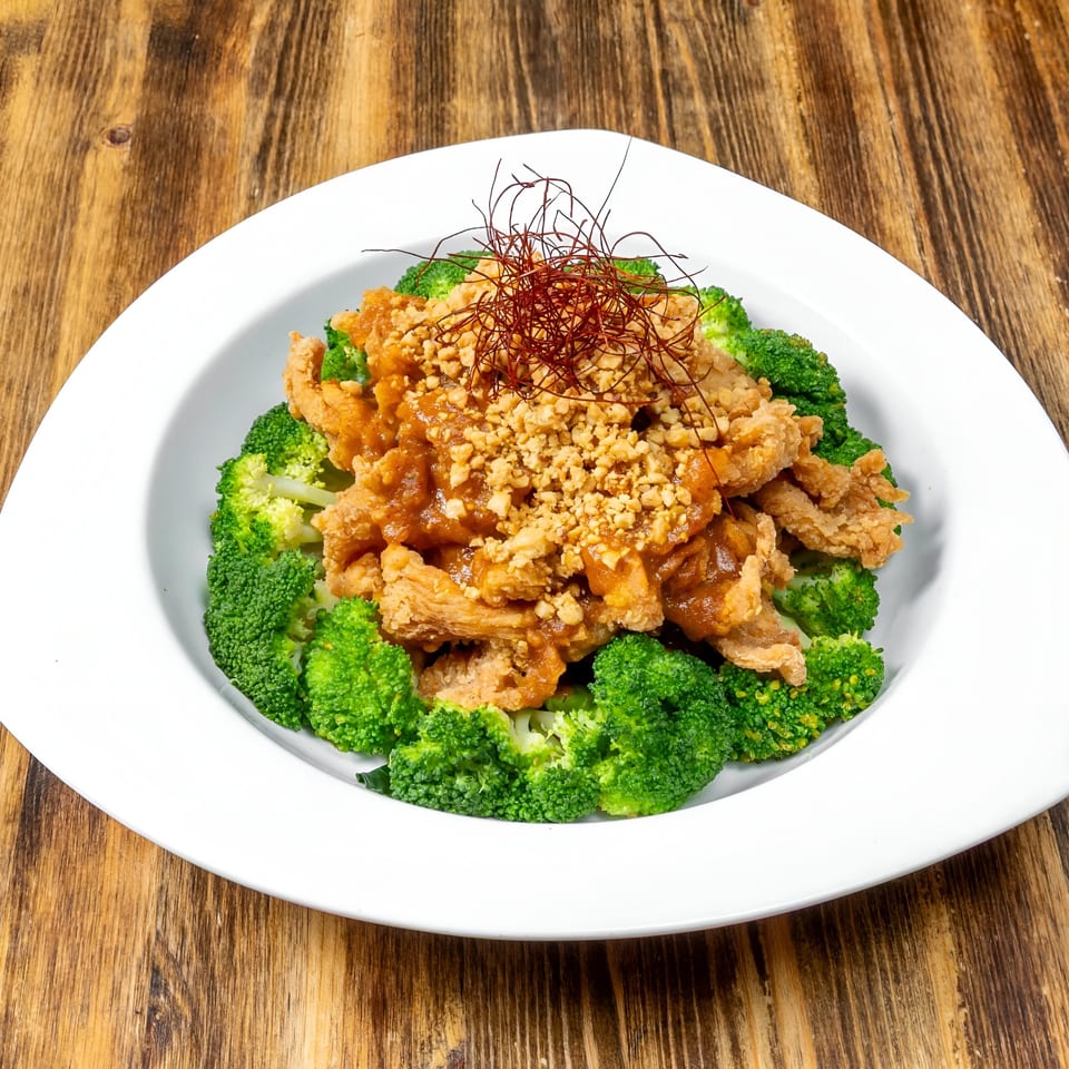 Crispy chicken with peanut sauce and chili threads, surrounded by steamed broccoli florets on a white plate.